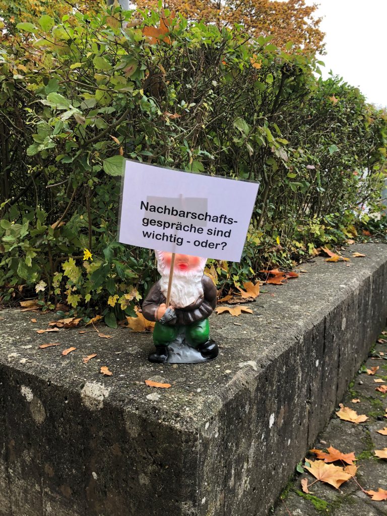 Zu erkennen ist ein kleiner Gartenzwerg, der ein kleines Plakat mit der Aufschrift "Nachbarschaftsgespräche sind wichtig - oder?" in die Hand hält. Aufgenommen wurde das Bild im Rahmen eines Projektbesuches in Heilbronn-Böckingen.