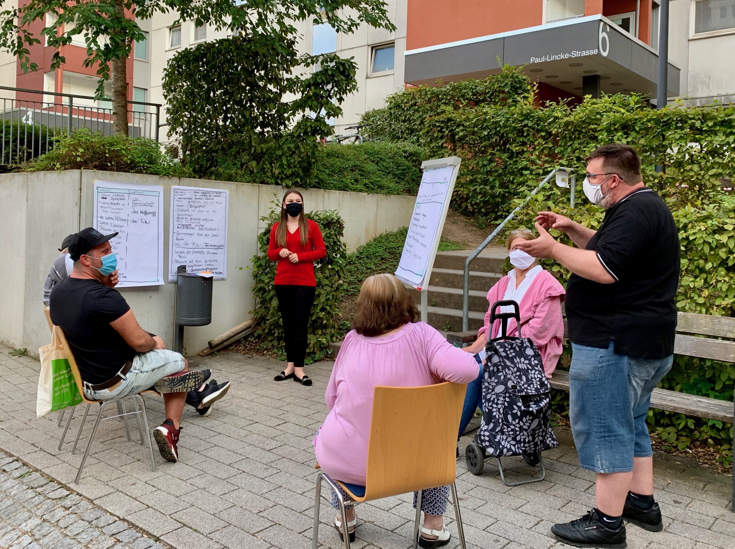 Zu sehen sind sechs Menschen, die auf einer Außenfläche vor einem Wohngebäude zusammengekommen sind und miteinander interagieren. Einige der Personen sitzen dabei auf Stühlen, während eine Person auf einer Bank sitzt und zwei Personen stehen. An der Außenfassade sowie vor dem Treppenaufstieg zum Wohngebäude wurden Flipcharts angebracht. Aufgenommen wurde das Bild im Rahmen der Nachbarschaftsgespräche in der Paul-Lincke-Straße in Stuttgart Botnang.