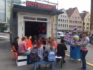 Zu sehen sind mehrere Menschen, die im Freien zusammengekommen sind und vor einem Container mit der Aufschrift "Botschaft" sitzen. Aufgenommen wurde das Bild im Rahmen der Nachbarschaftsgespräche in Ulm.