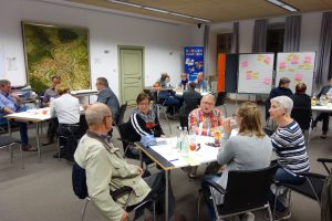 Zu sehen sind eine Vielzahl an Menschen, die in einem großen Raum zusammengekommen sind und an mehreren Tischen verteilt sitzen und miteinander sprechen. Entstanden ist das Bild im Rahmen eines Nachbarschaftsgespräches in Sulz am Neckar.