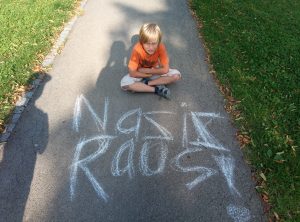 Zu sehen ist ein kleiner Junge, der auf dem Gehweg sitzt und in die Kamera blickt. Auf dem Gehweg steht die Aufschrift "Nazis raus" Aufgenommen wurde das Bild im Rahmen eines Projektbesuches des "Guter Draht Esel" Projektes, gefördert durch das Förderprogramm Nachbarschaftsgespräche.
