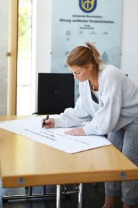 Zu sehen ist eine Frau, die etwas auf ein großes weißes Plakat schreibt. Entstanden ist das Bild im Rahmen eines Nachbarschaftsgespräches.